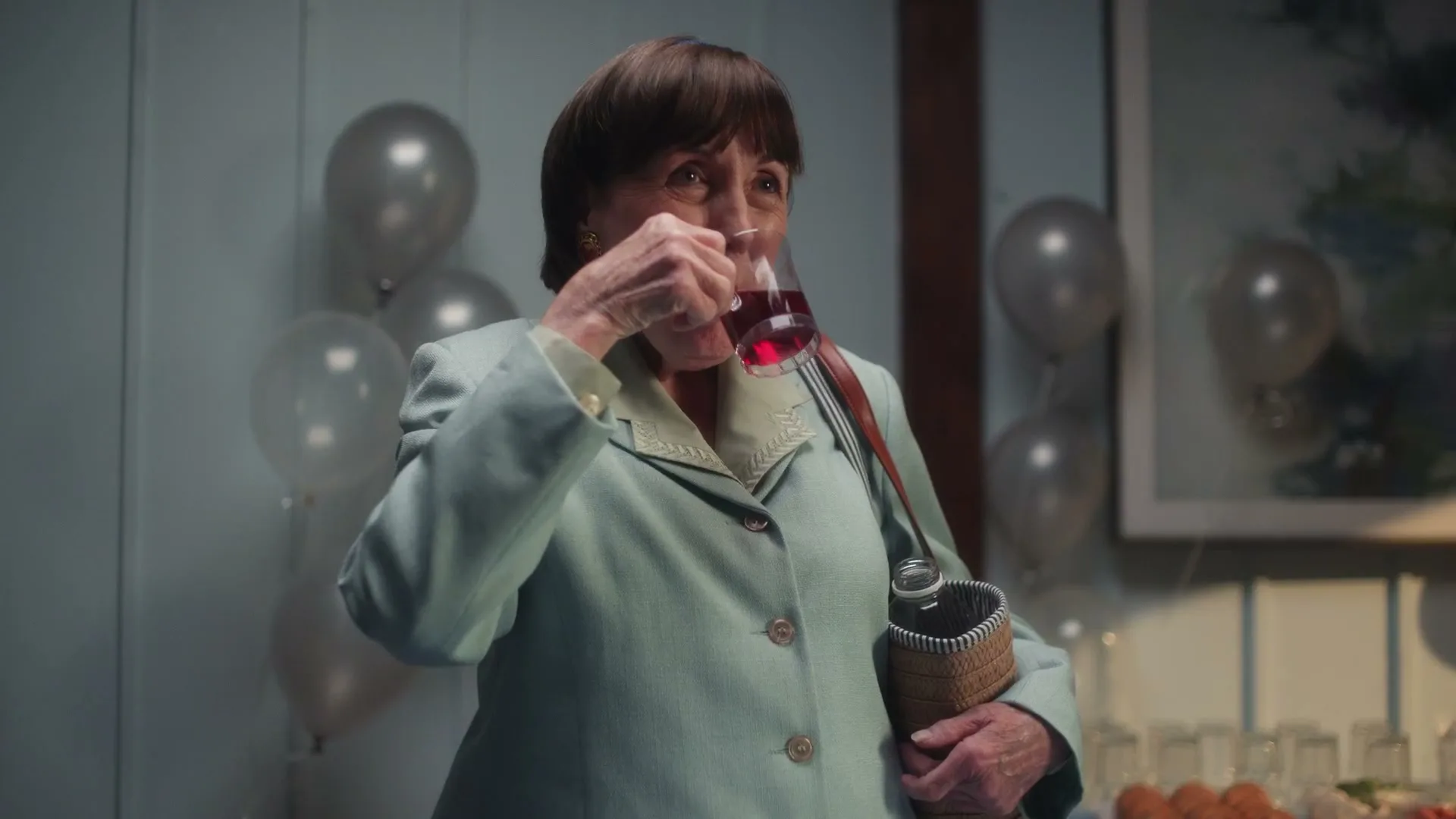 An older woman in a light green suit sips a drink from a small glass, holding a woven bag, at an indoor party with balloons.
