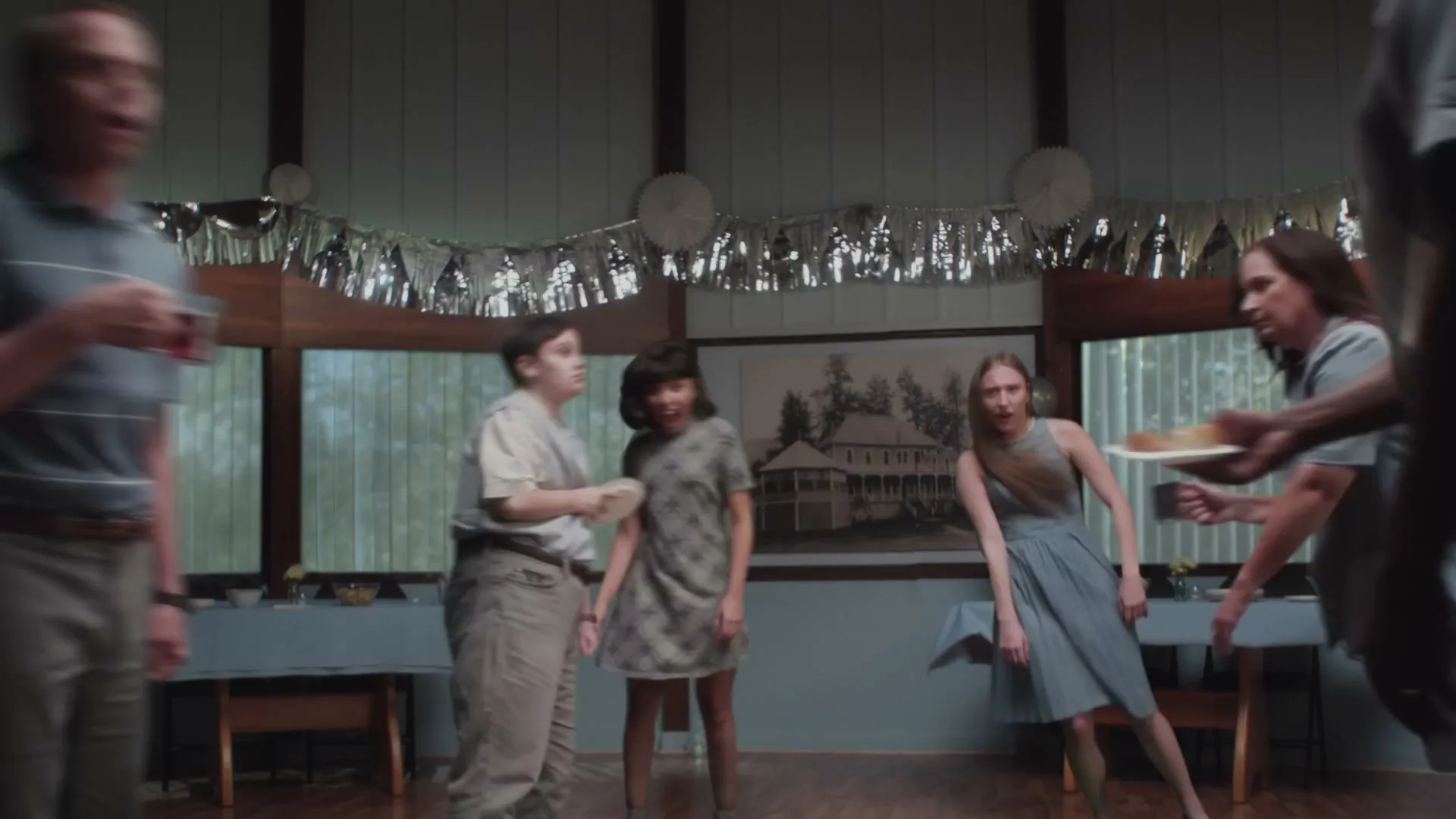 A group of people, including a man in a polo shirt, a boy in a button-up, and two women in dresses, are dancing awkwardly at a party in a house.