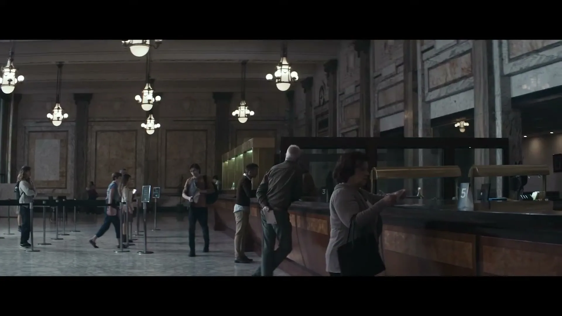 A diverse group of people, dressed in casual attire, are seen waiting in line and interacting with tellers at a grand, old-fashioned bank.