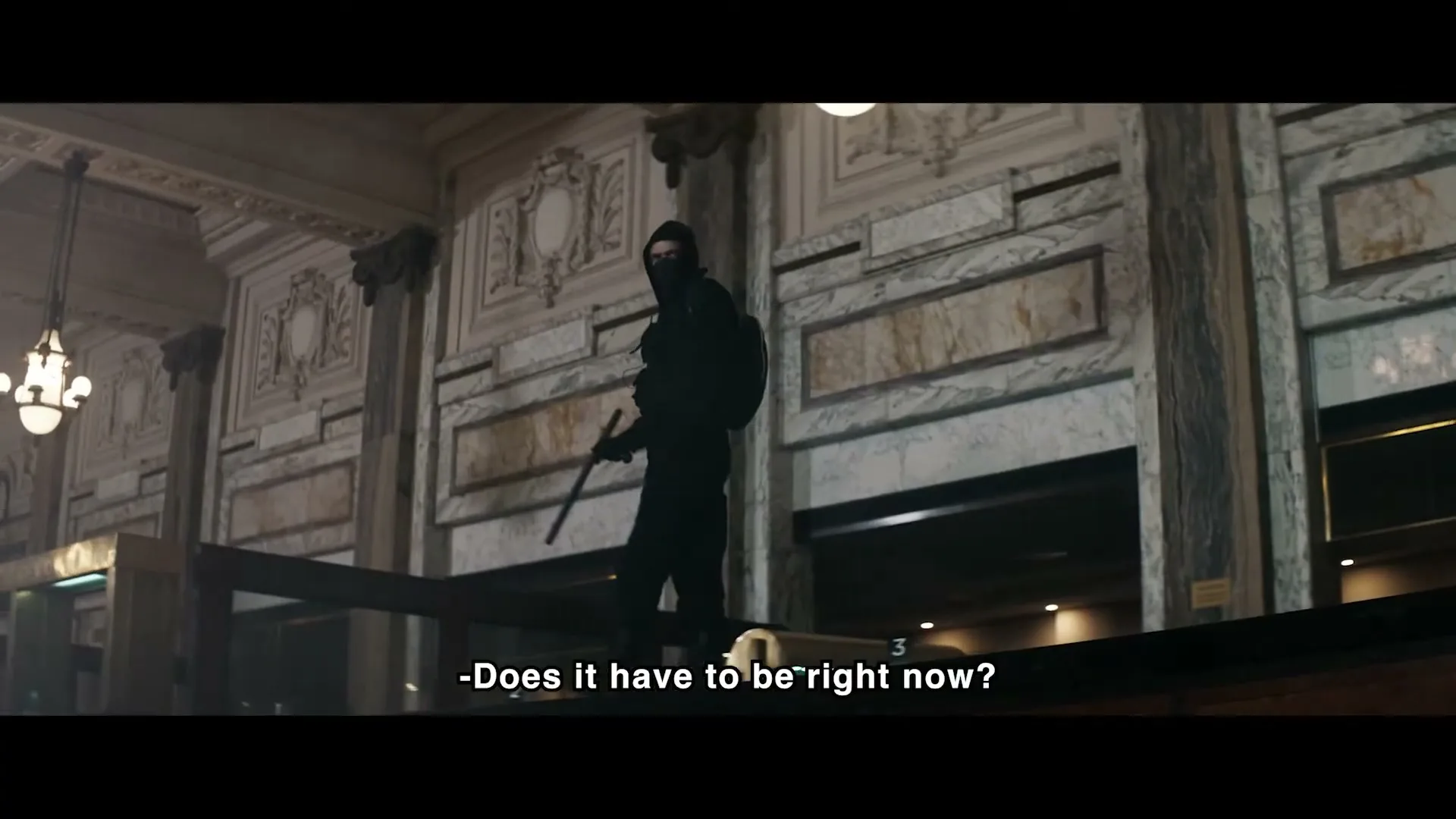 A person in a black hoodie and mask stands on a ledge in a grand, ornate building, looking down, with subtitles visible.