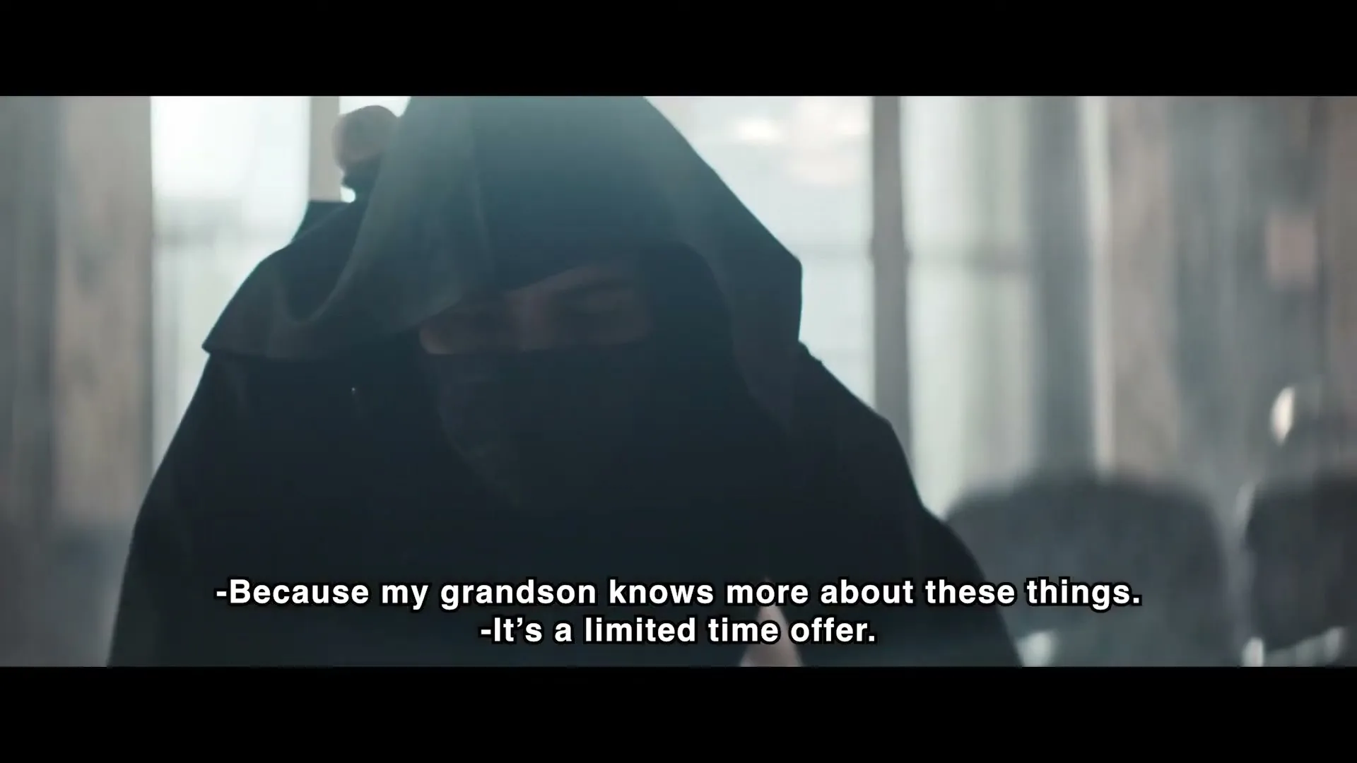 A person in a dark hooded cloak and face covering looks down, then up, in a dimly lit interior space.