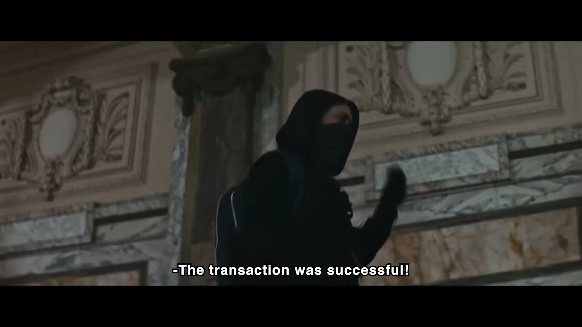 A person in a black hoodie and face covering stands in a grand, ornate hall, gesturing with their hands as if speaking to someone off-screen.