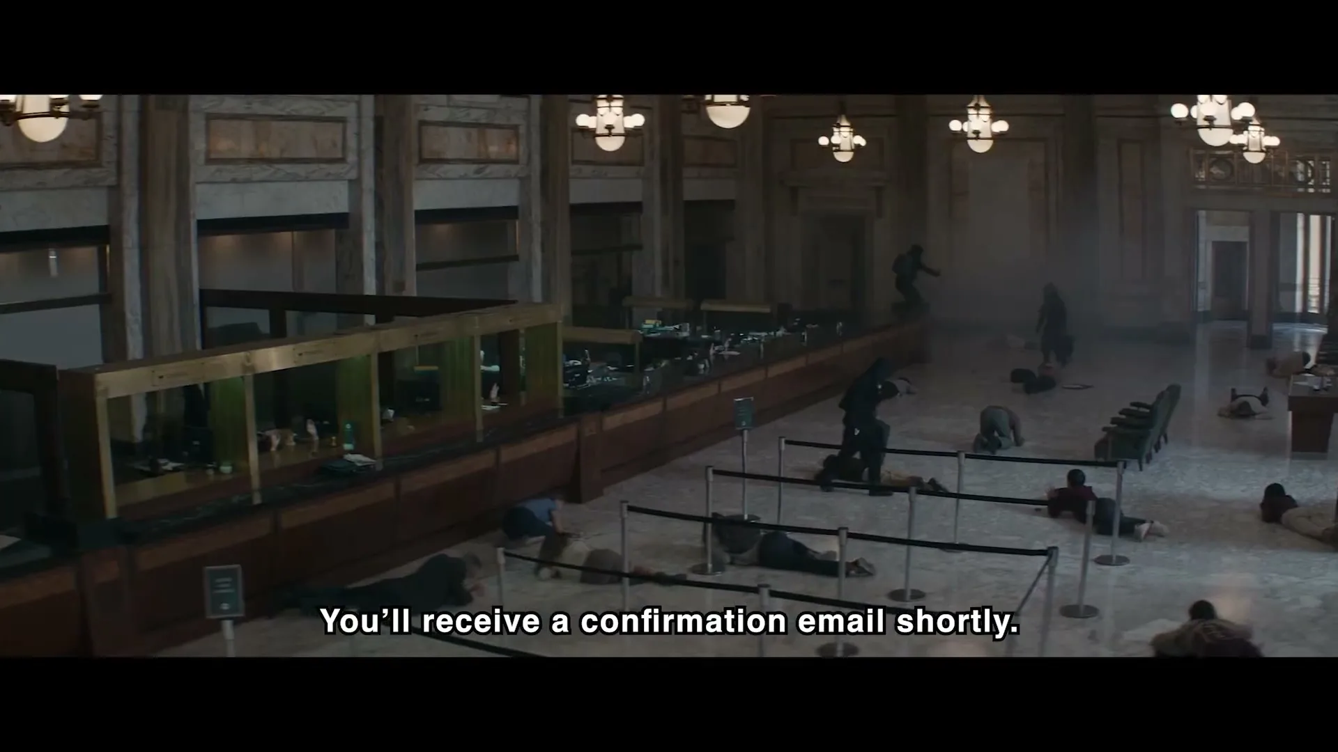 A wide shot shows a bank interior with many people lying on the floor, some crawling, as a person in dark clothing walks over them.