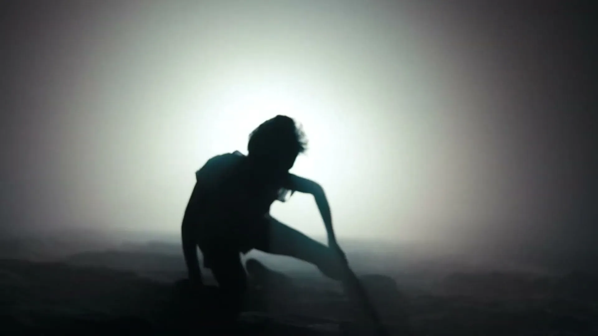 A child's silhouette is seen against a bright, hazy light, slowly collapsing to the ground in a desolate, foggy landscape.