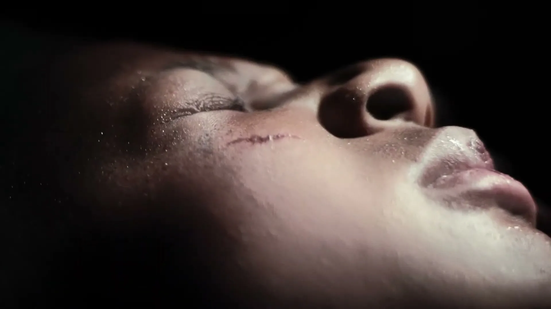 A person's face with a scar on their cheek is shown in a close-up, with dramatic lighting highlighting their features against a dark background.
