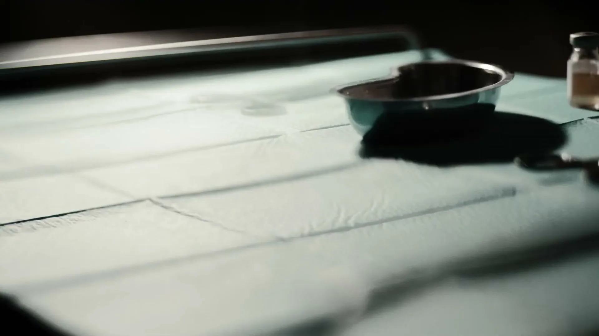 A close-up shot shows medical instruments, including scissors and a kidney dish, being placed on a sterile green sheet on a table in a dimly lit room.