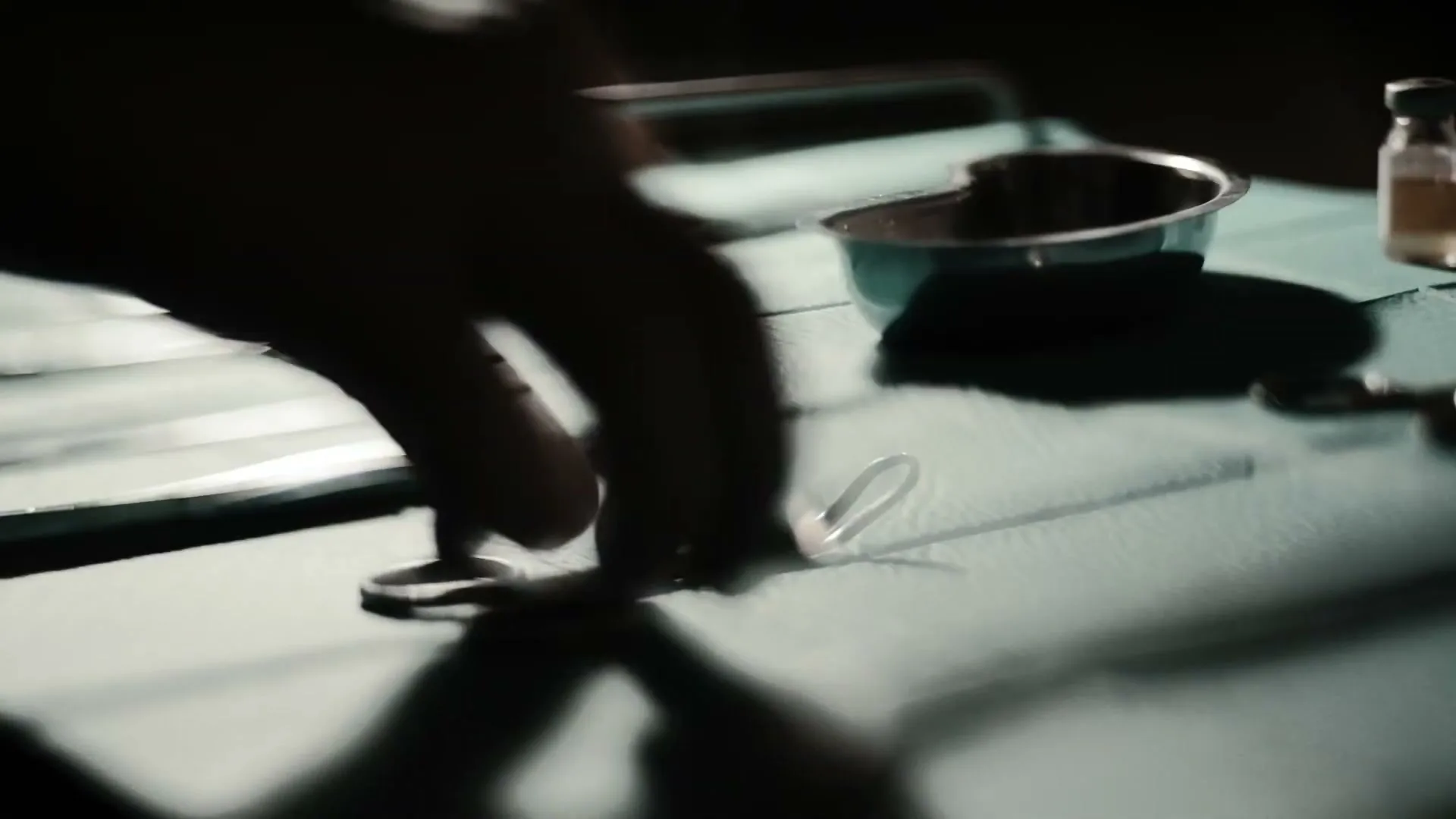 A surgeon's hands are silhouetted as they pick up a surgical clamp from a sterile green cloth, with other instruments nearby.