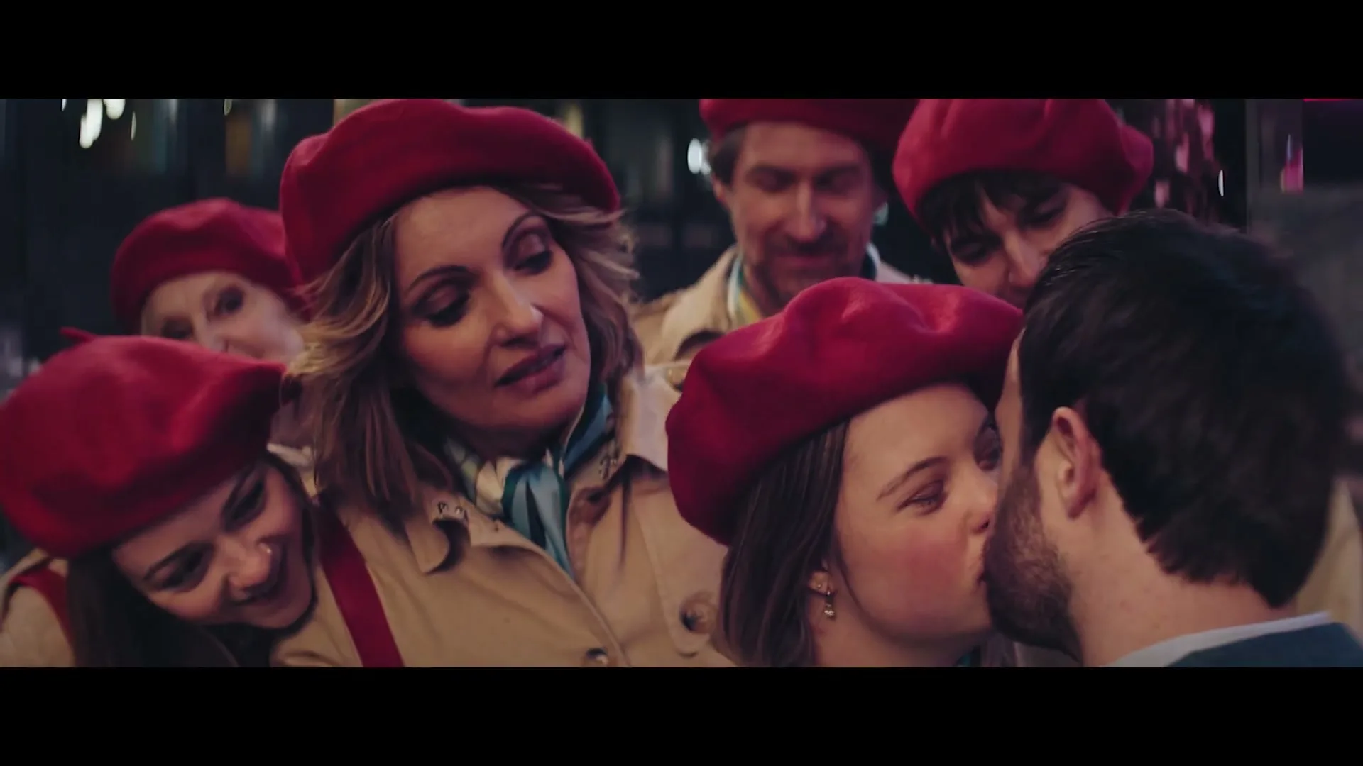 A young woman with Down syndrome kisses a man, surrounded by a group of people in red berets and trench coats, on a city street at night.