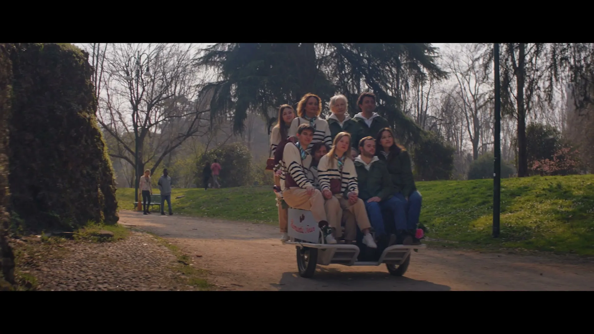 A group of young people, some wearing matching striped sweaters, are riding together on a small, open-air vehicle down a path in a park on a sunny day.