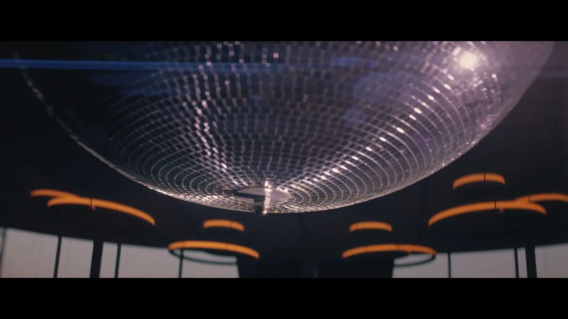 A large, sparkling disco ball hangs from the ceiling, reflecting light in a dark room with orange-lit circular fixtures.