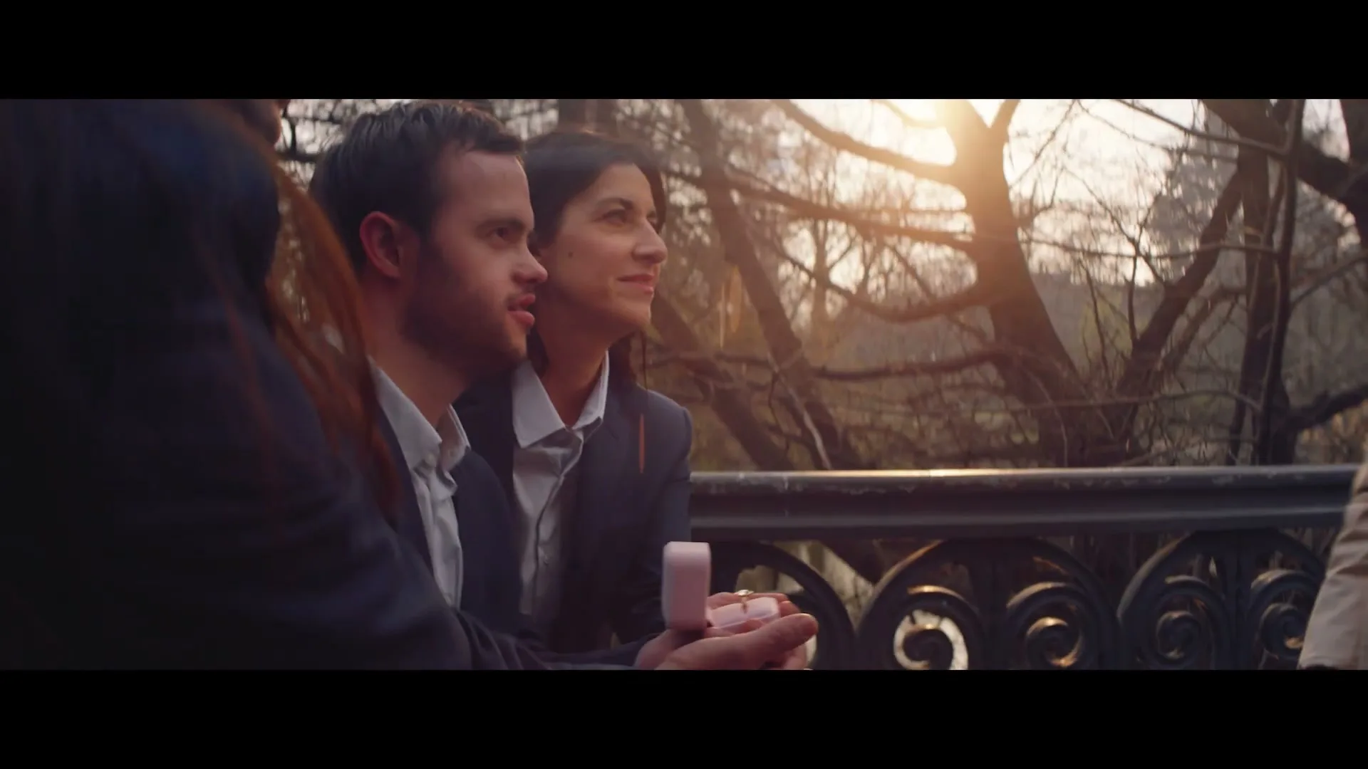 A man with Down syndrome proposes to a woman with a pink ring box on a bridge in a park at golden hour.