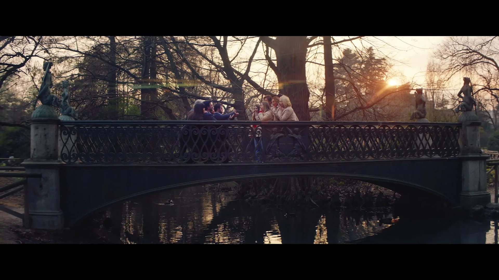 A group of friends, including a woman with Down syndrome, stand on a bridge in a park at sunset, taking a selfie.