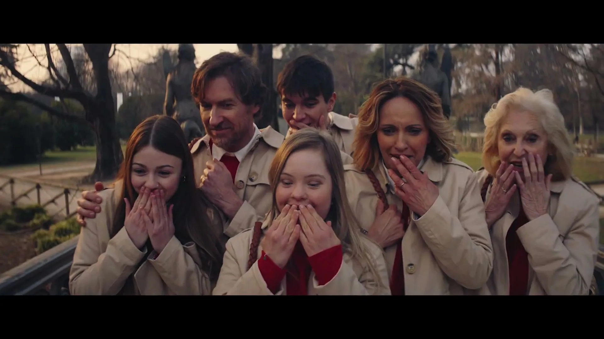 A diverse group of six people, including a young woman with Down syndrome, stand together in a park, all wearing matching trench coats and red shirts, covering their mouths in surprise.
