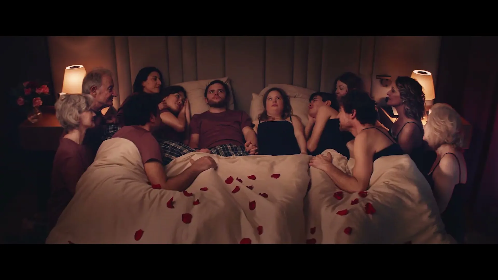 A couple with Down syndrome lies in bed, holding hands, surrounded by many people of various ages, all looking at them in a dimly lit bedroom.