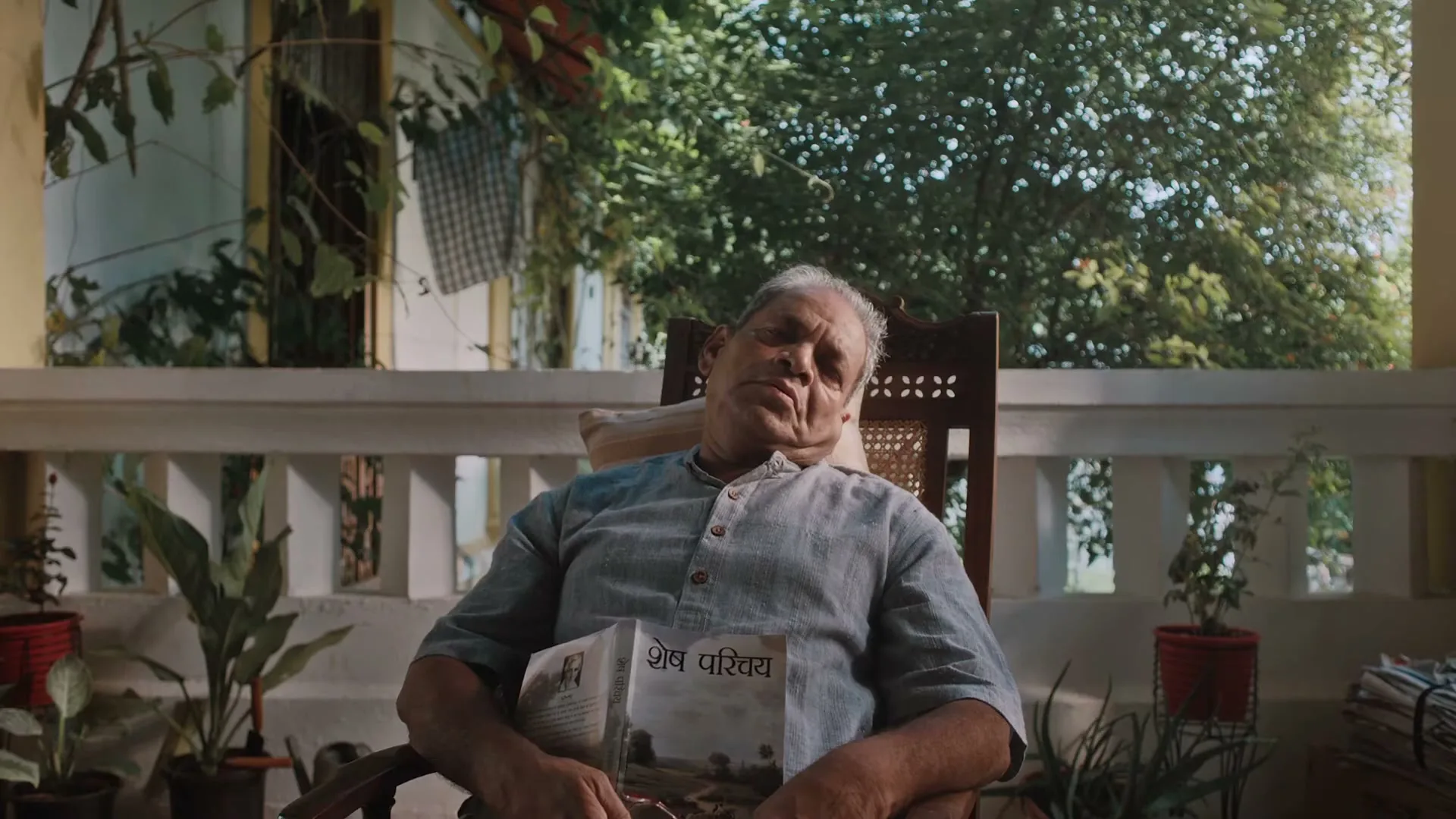 An elderly man in a light blue shirt is sleeping in a wooden chair on a porch, holding a book, then wakes up suddenly.