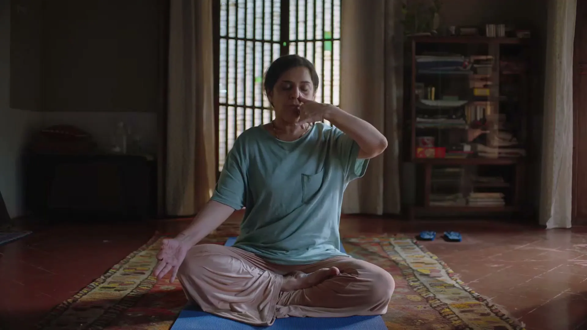 A woman in her 40s practices alternate nostril breathing on a yoga mat in a dimly lit room, with sunlight filtering through a window.