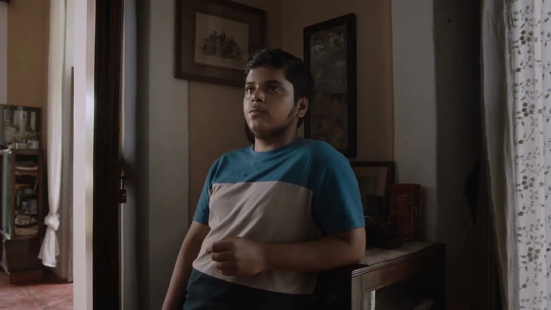 A young boy with a striped t-shirt leans on a wooden table, looking up and to the right with a surprised expression in a dimly lit room.
