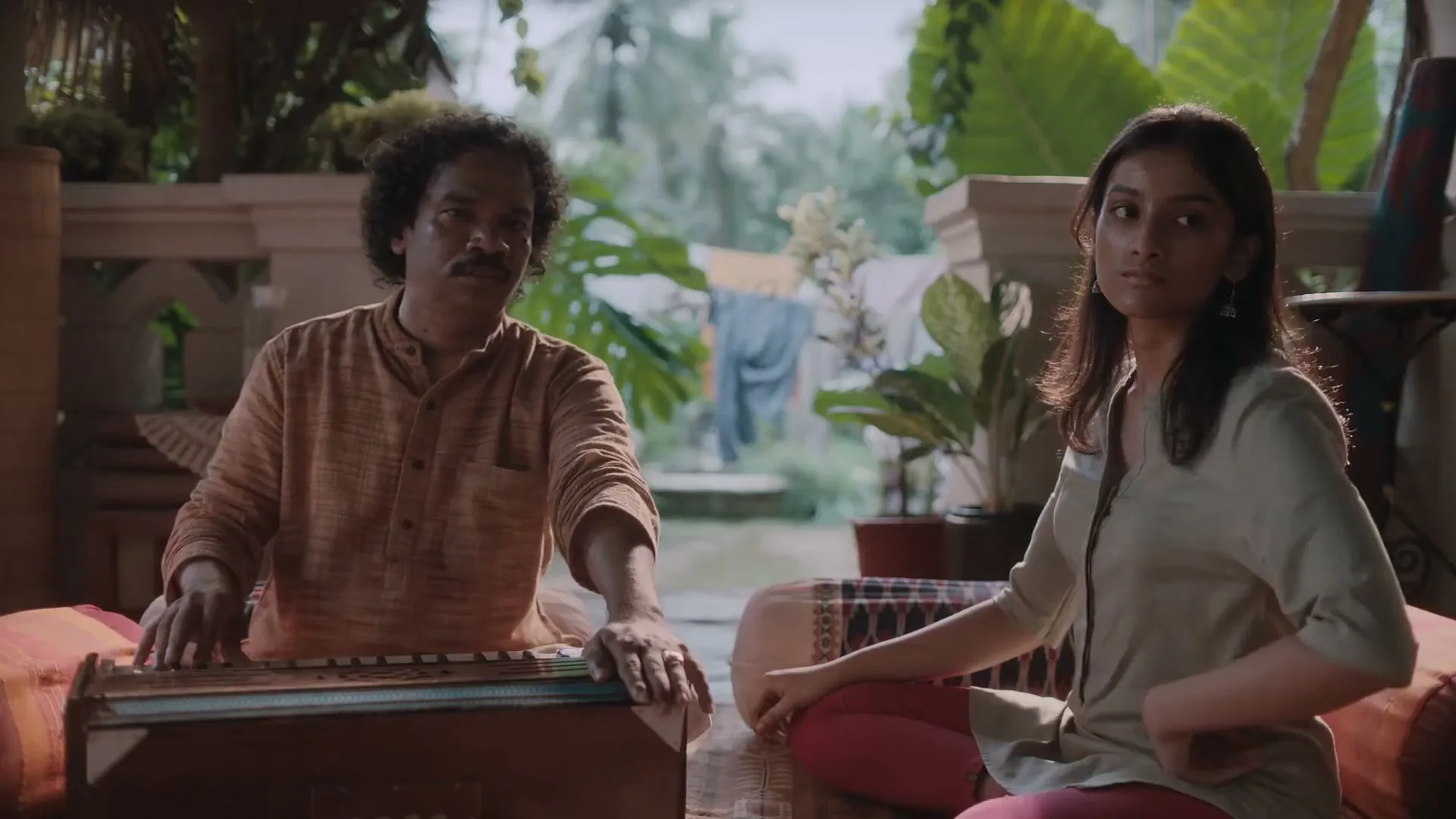 A man with curly hair plays a harmonium while a young woman sits beside him, both looking at the camera on a porch.