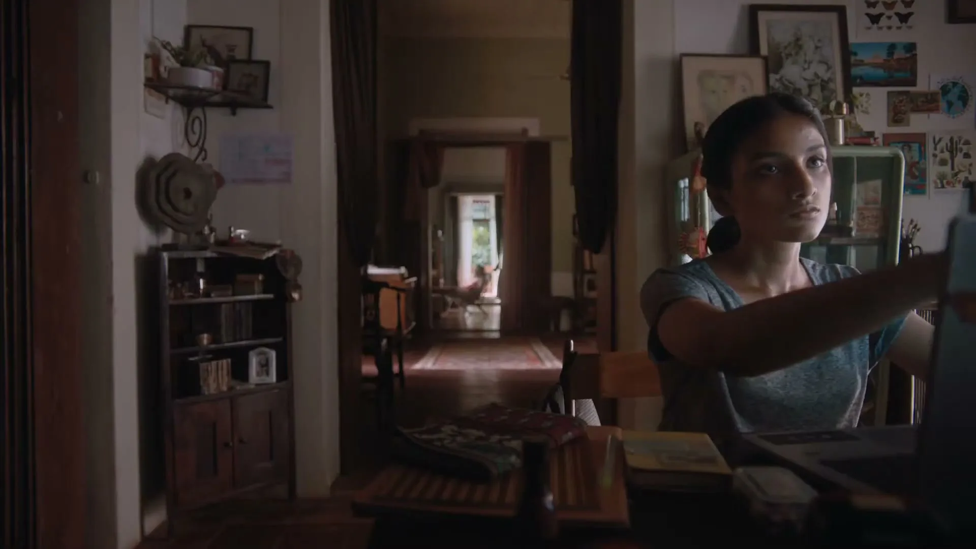 A young woman with dark hair in a ponytail sits at a wooden desk, looking intently at a laptop screen in a dimly lit room.
