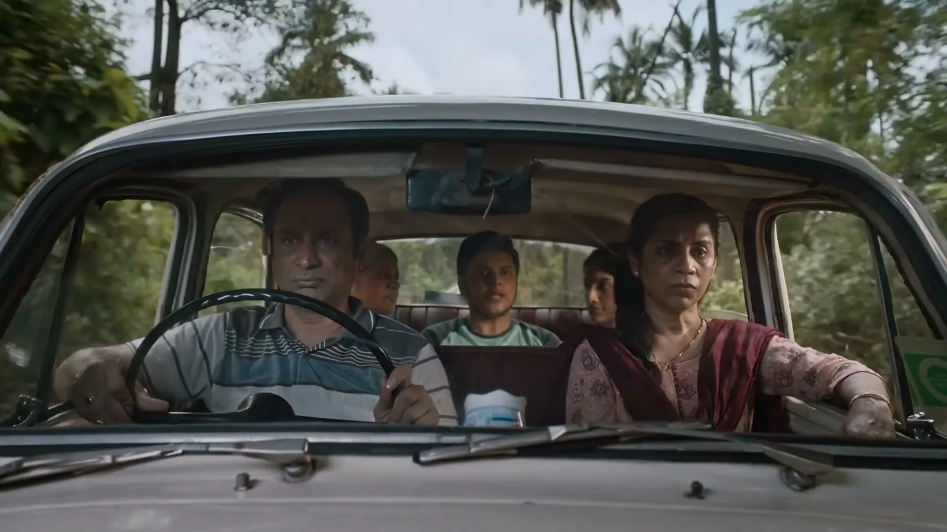 A family of four, including a man driving and a woman in the passenger seat, look ahead with serious expressions while driving in a car on a tree-lined road.