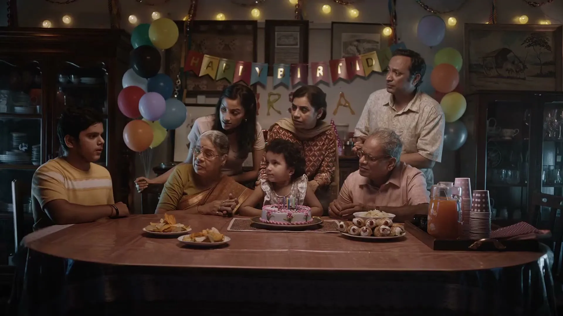 A multi-generational Indian family gathers around a table to celebrate a young girl's birthday, with balloons and a "Happy Birthday" banner in the background.