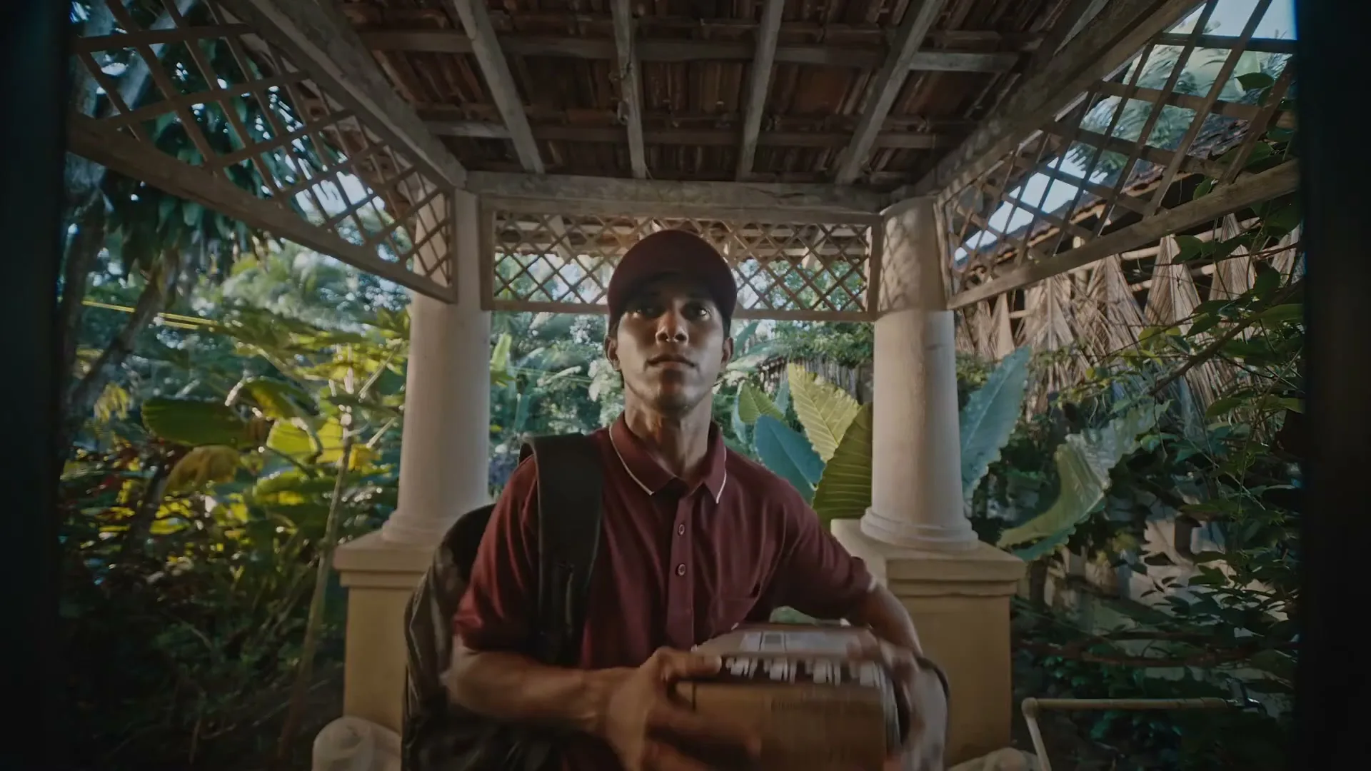 A young delivery man in a red uniform and cap holds a package, looking up with a serious expression under a wooden pergola.