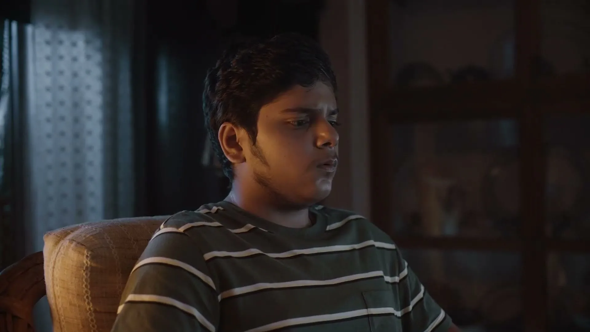 A young man with dark hair and a striped t-shirt sits in a chair, looking intently at something off-screen in a dimly lit room.