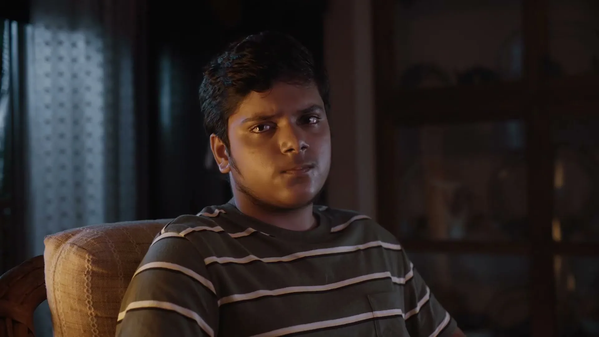A young male teen with dark hair and a striped t-shirt sits in a chair, looking forward with a neutral expression in a dimly lit room.