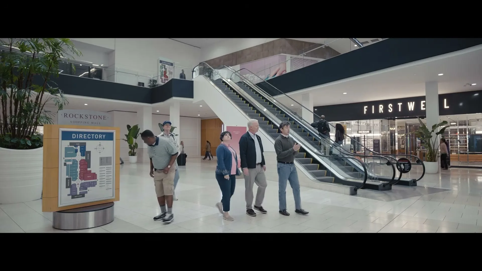 A diverse group of adults walks through a bright, modern shopping mall, with one man looking at a directory and another checking his watch.