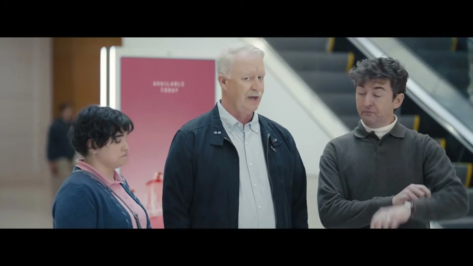 A man in a dark sweater checks his watch while two other people, a woman and an older man, stand beside him in a brightly lit mall.