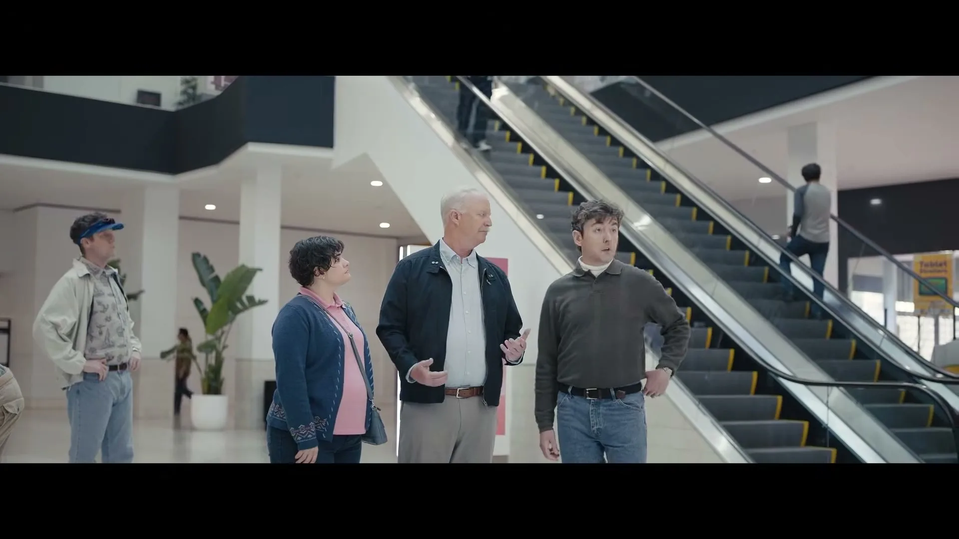 Dr. Rick, a middle-aged man in a jacket, talks to a small group of young homeowners in a brightly lit mall atrium with escalators in the background.
