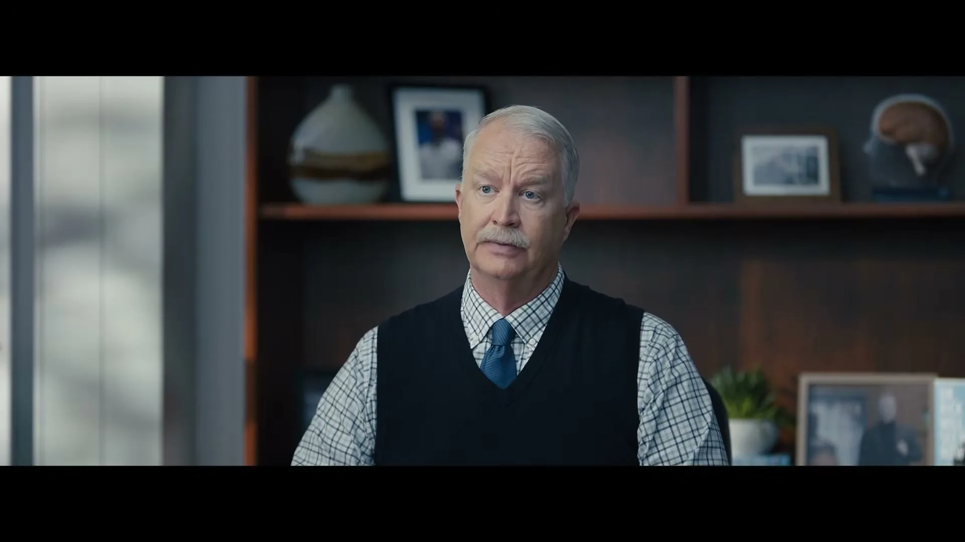 A male therapist with a mustache and gray hair, wearing a sweater vest and tie, sits in his office, looking directly at the camera.