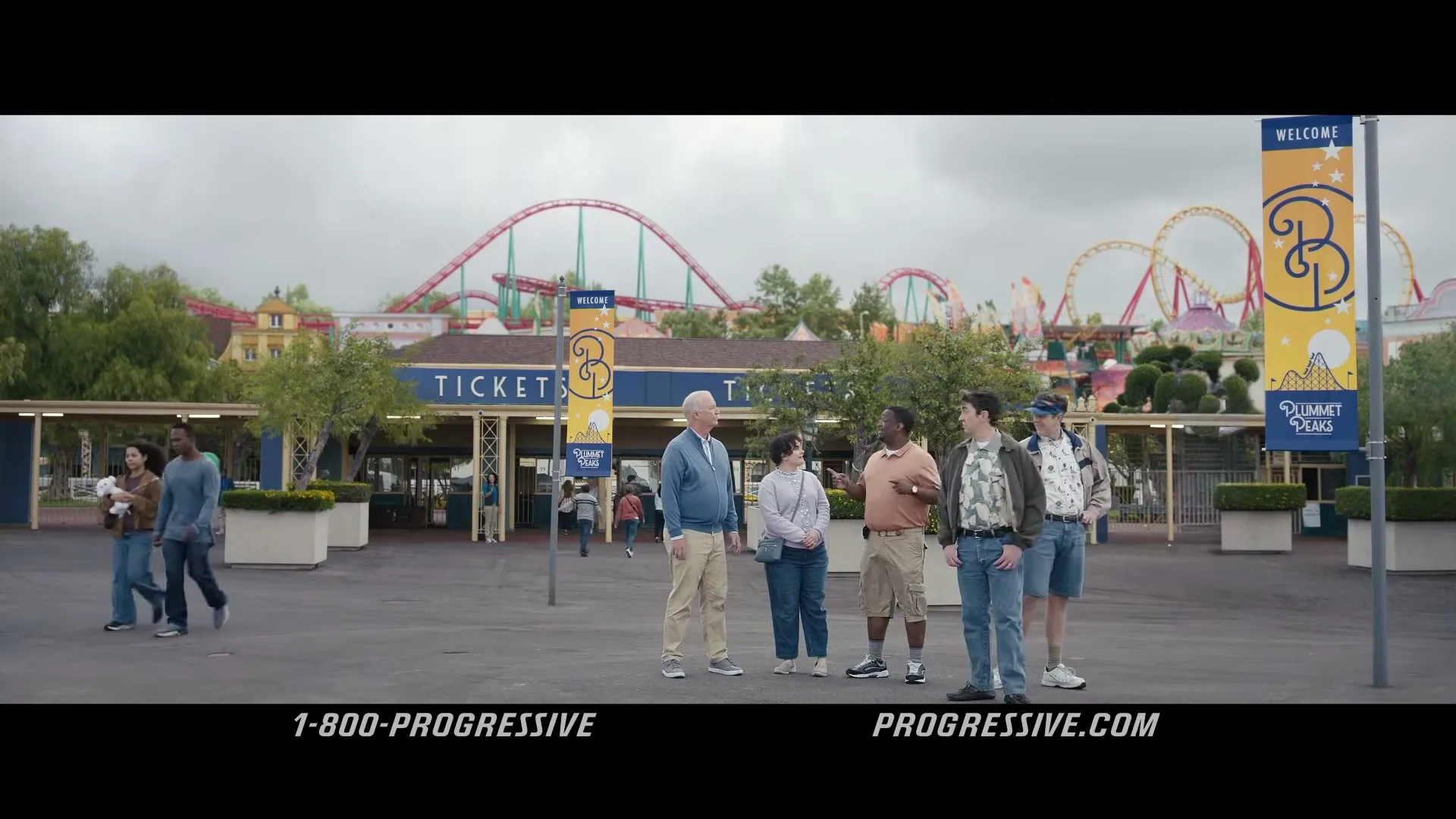A group of five adults stands in front of a theme park entrance, with roller coasters visible in the background under a cloudy sky.