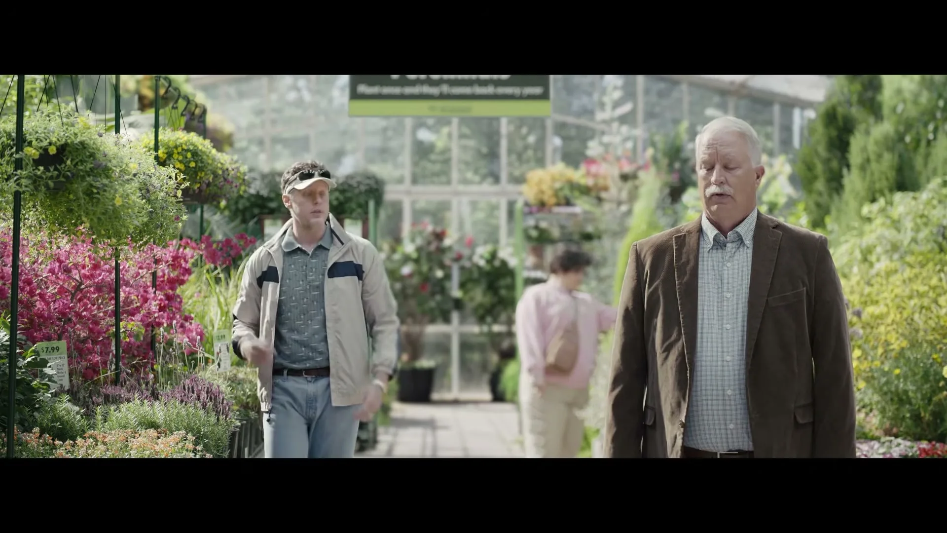 A man in a visor and a man in a brown jacket stand in a plant nursery, looking around with confused expressions.