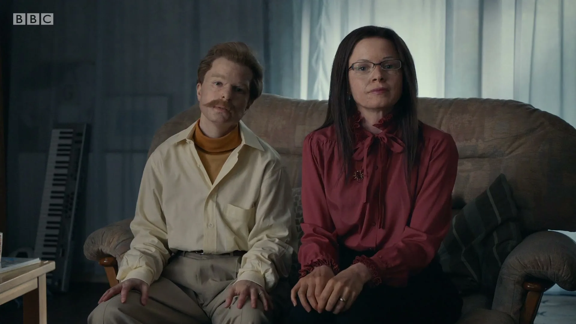 A middle-aged couple, dressed in retro clothing, sits on a sofa in a dimly lit living room, looking directly at the camera with serious expressions.