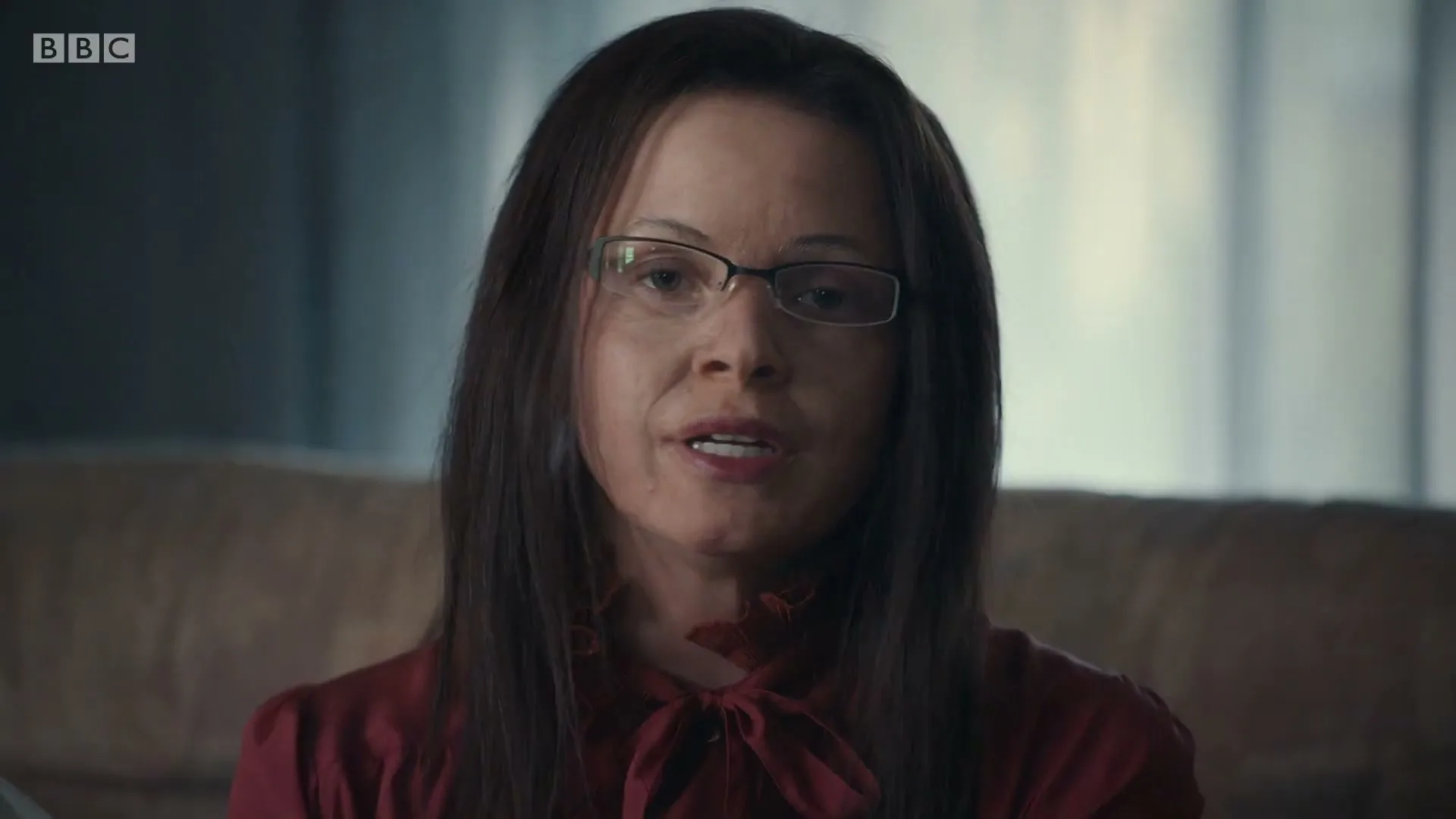 A woman with long dark hair and glasses, wearing a red blouse, looks directly at the camera in an interior setting.