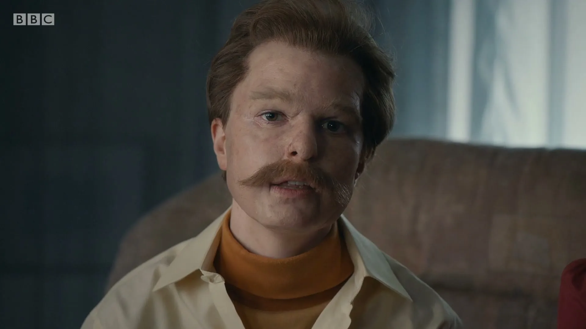 A man with a fake mustache and wig, wearing a collared shirt and turtleneck, looks around suspiciously in a dimly lit room.