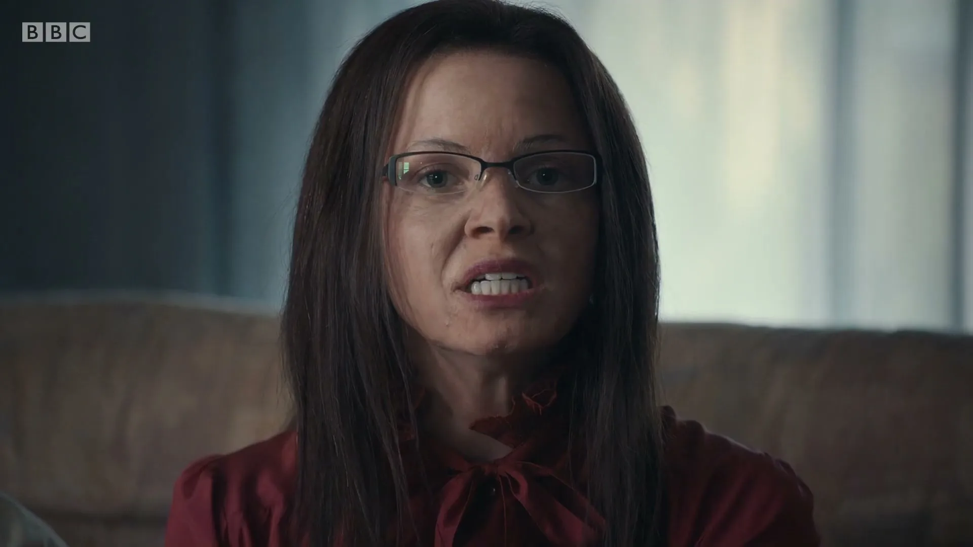 A woman with long brown hair and glasses, wearing a red blouse, looks directly at the camera with a neutral expression in an interior setting.