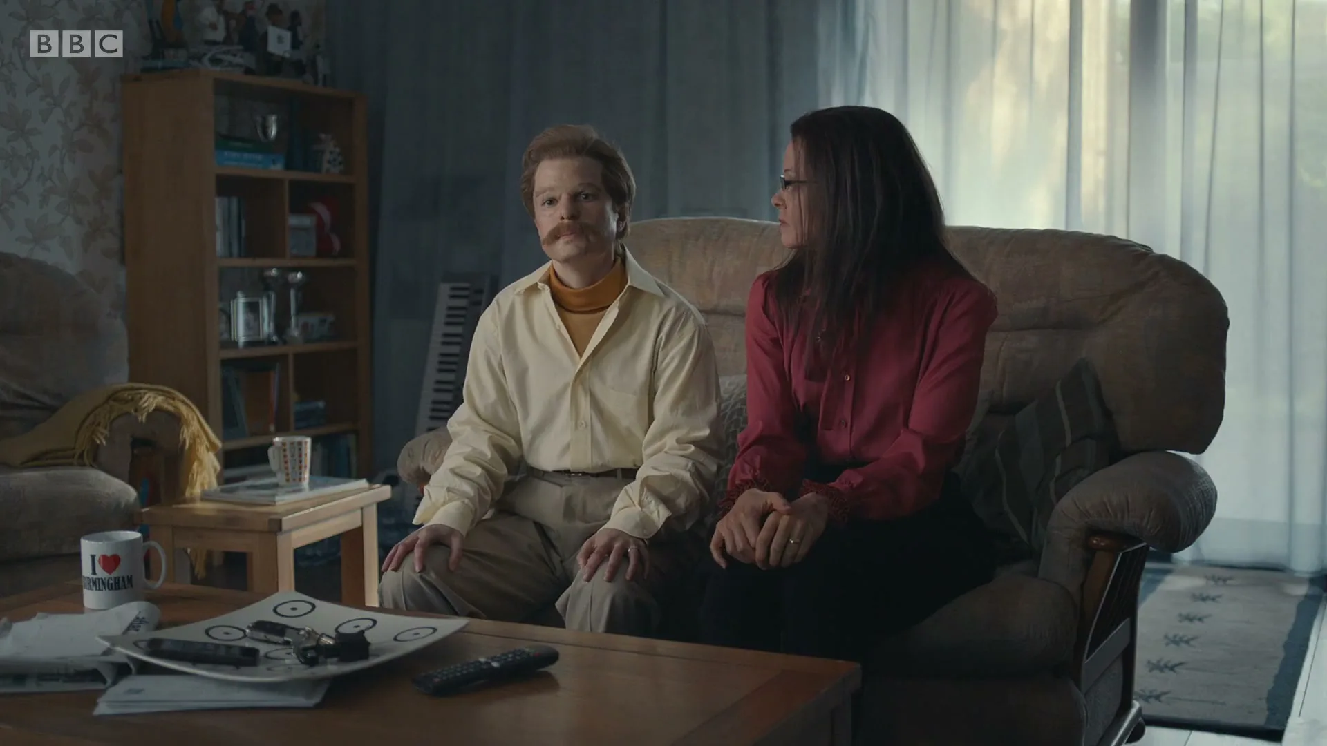 A man with a mustache and a woman with long dark hair sit on a couch in a living room, looking forward.