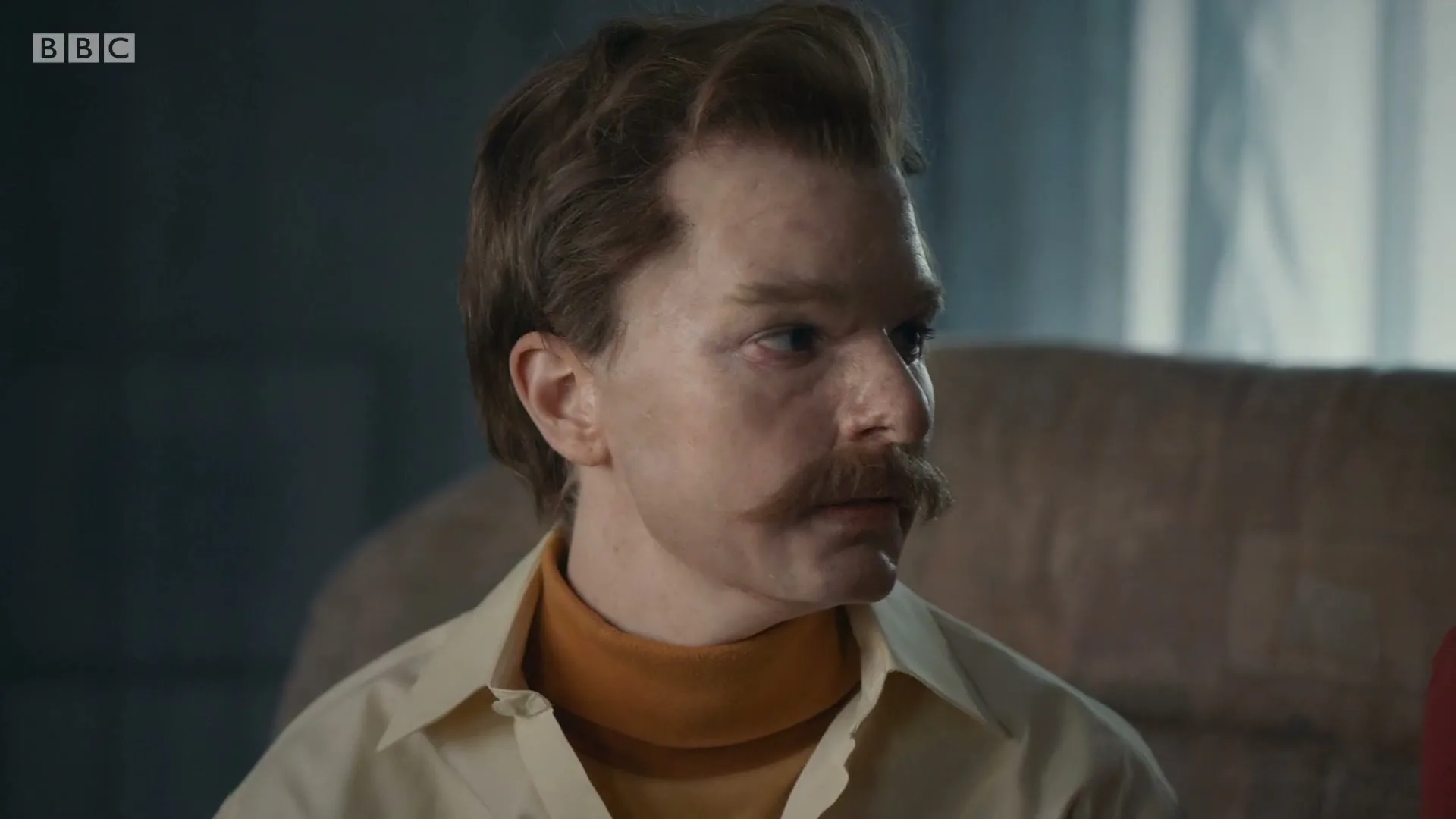 A man with a large mustache and a retro outfit looks around a dimly lit room, then directly at the camera.