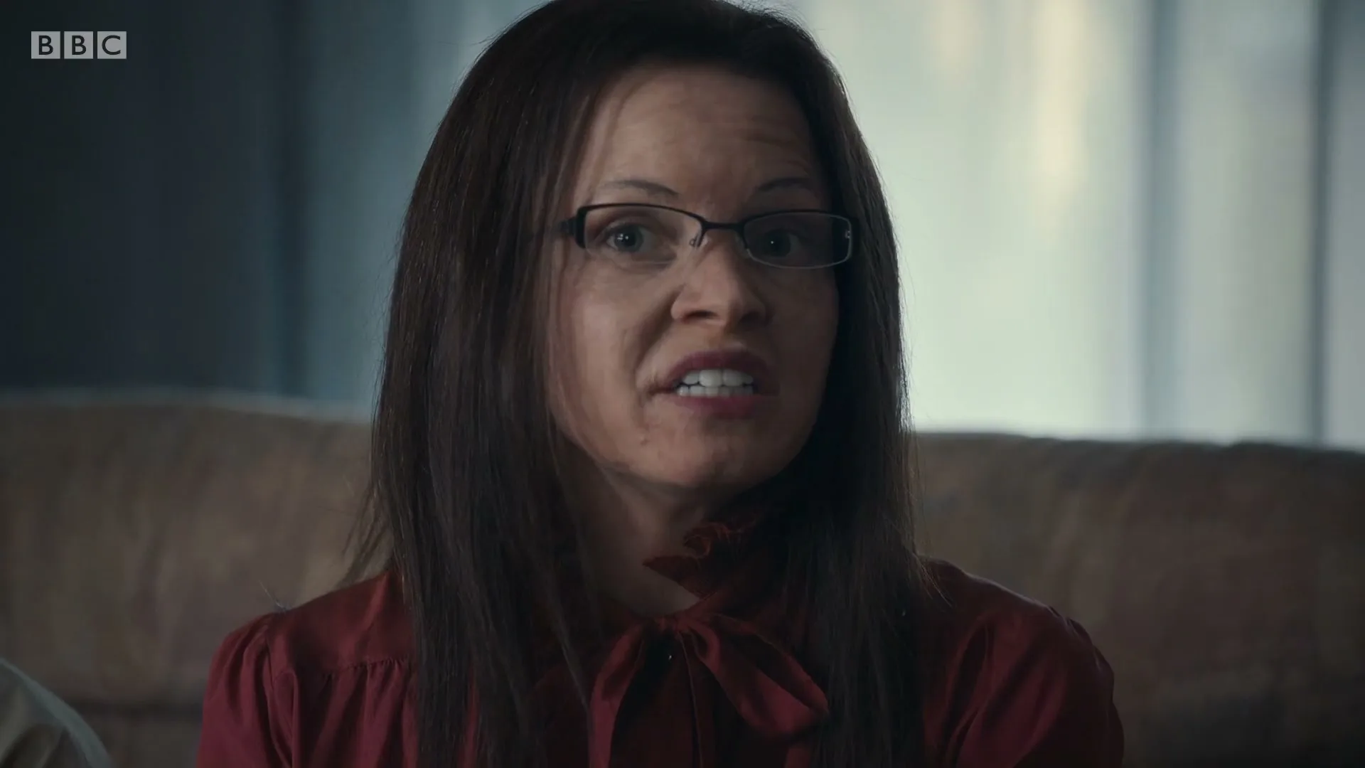 A woman with long dark hair and glasses, wearing a red blouse, sits on a couch and speaks in a living room.