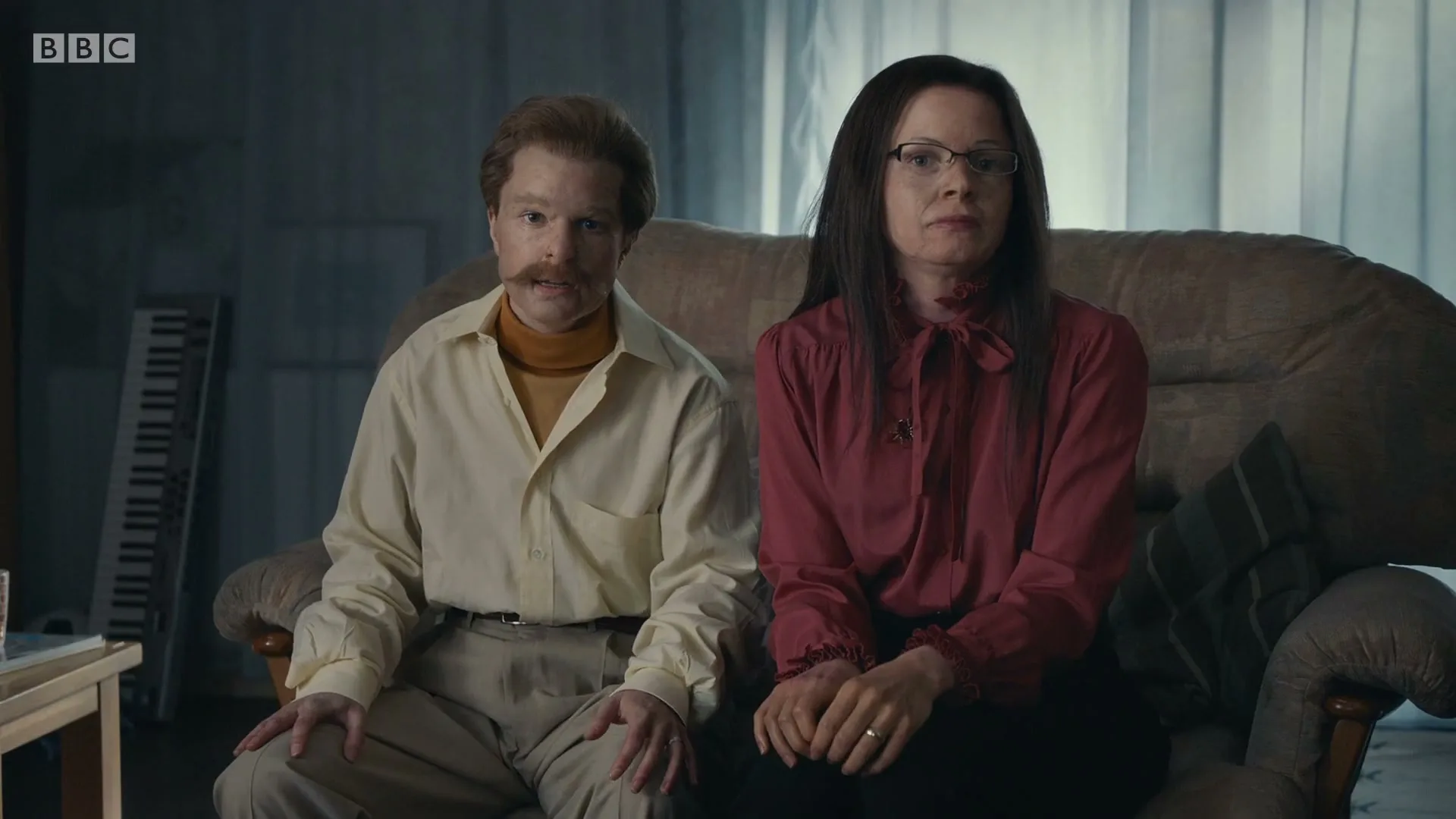 A man with a mustache and a woman with glasses sit on a couch, looking directly at the camera in a dimly lit room.