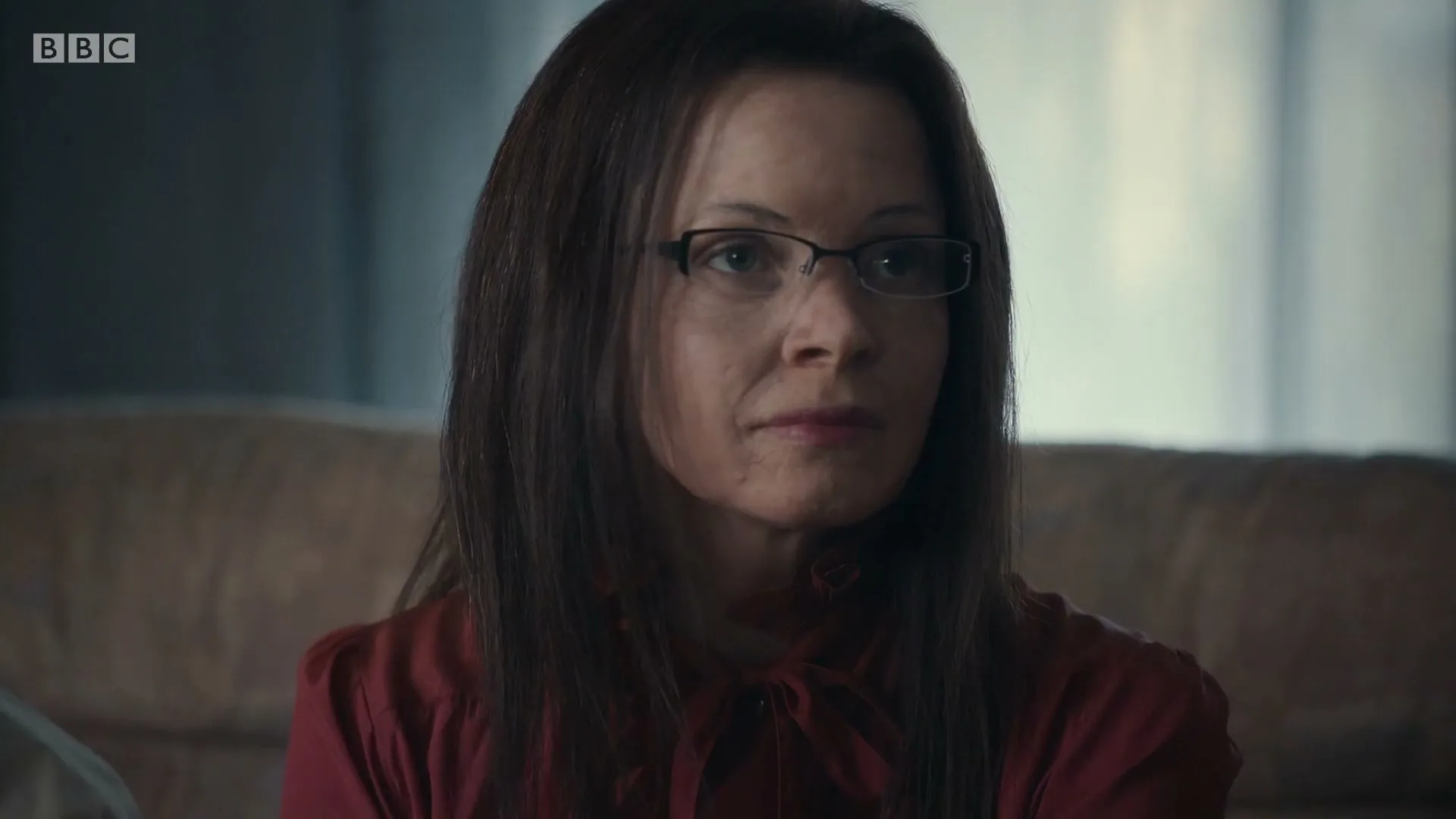 A woman with long brown hair and glasses, wearing a red blouse, looks directly at the camera with a neutral expression in an interior setting.
