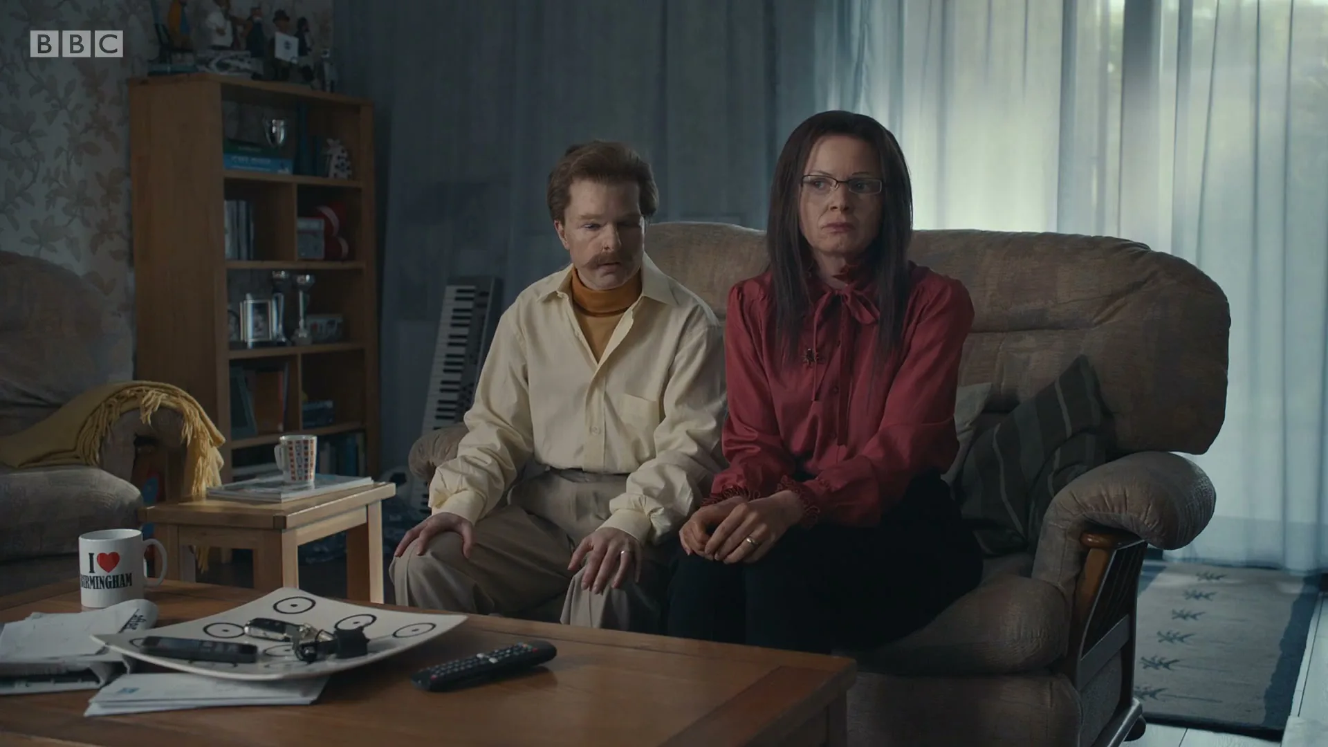 A man and a woman, dressed in retro clothing, sit side-by-side on a sofa in a living room, looking directly at the camera with serious expressions.