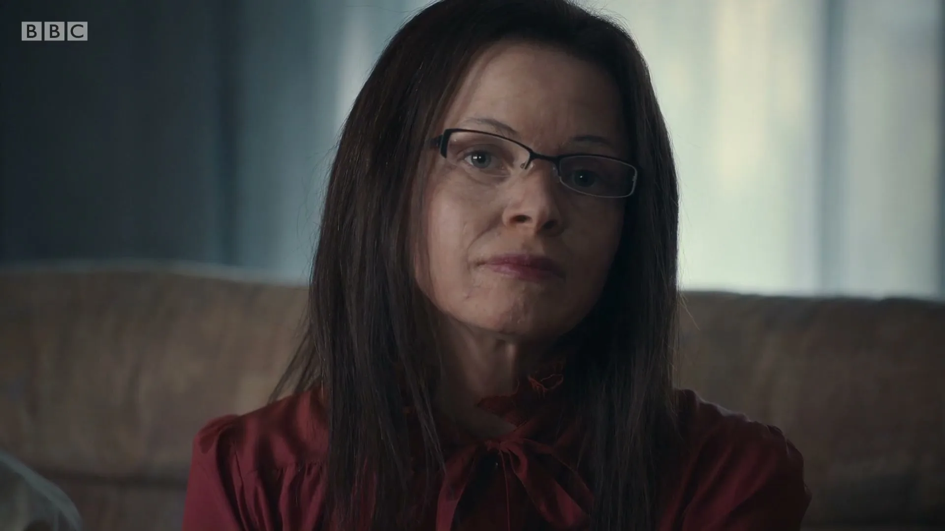A woman with long dark hair and glasses, wearing a red blouse, looks directly at the camera with a neutral expression in an interior setting.