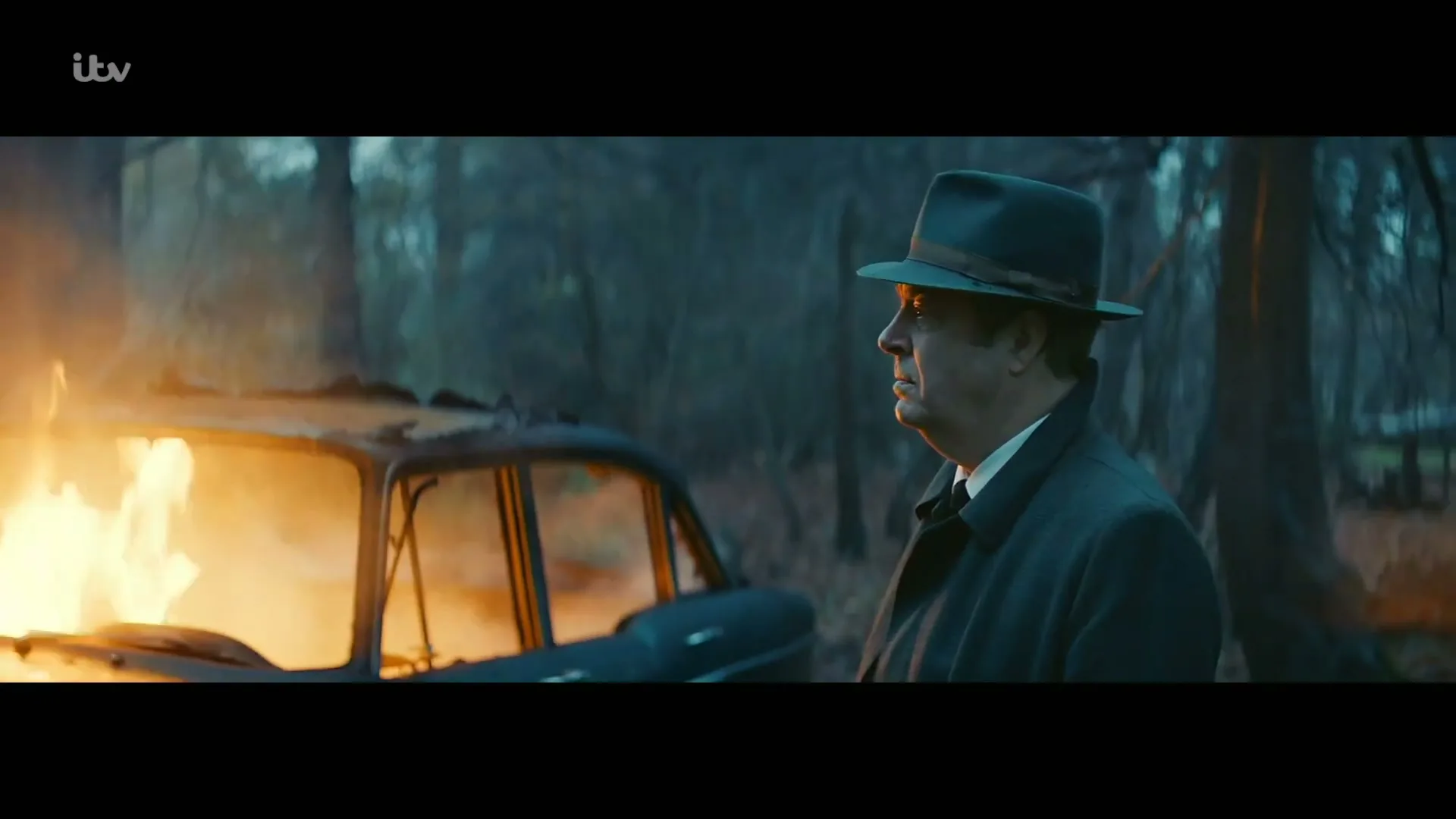 A man in a fedora and trench coat stands next to a burning car in a forest, looking off-screen with a serious expression.