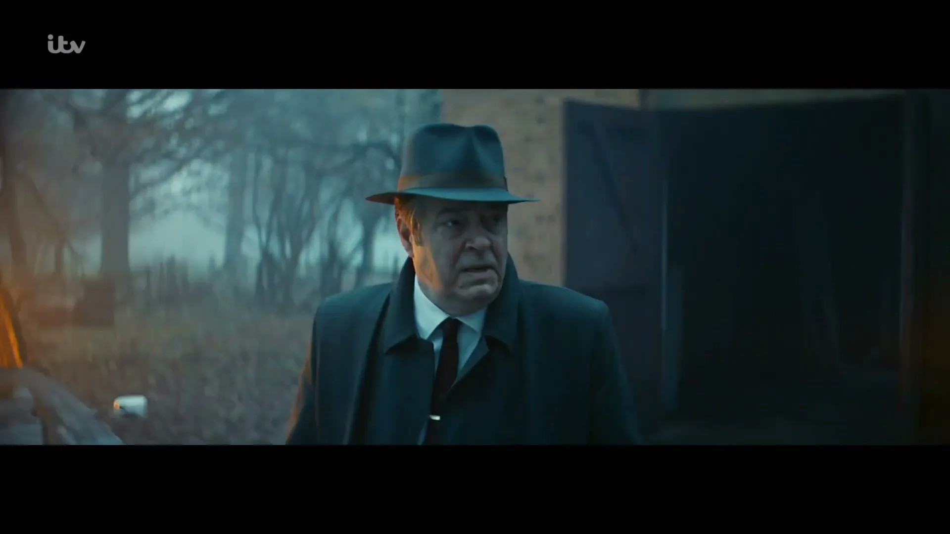 A man in a fedora and trench coat walks through a misty, dark landscape with bare trees and a brick building in the background, looking directly at the camera.
