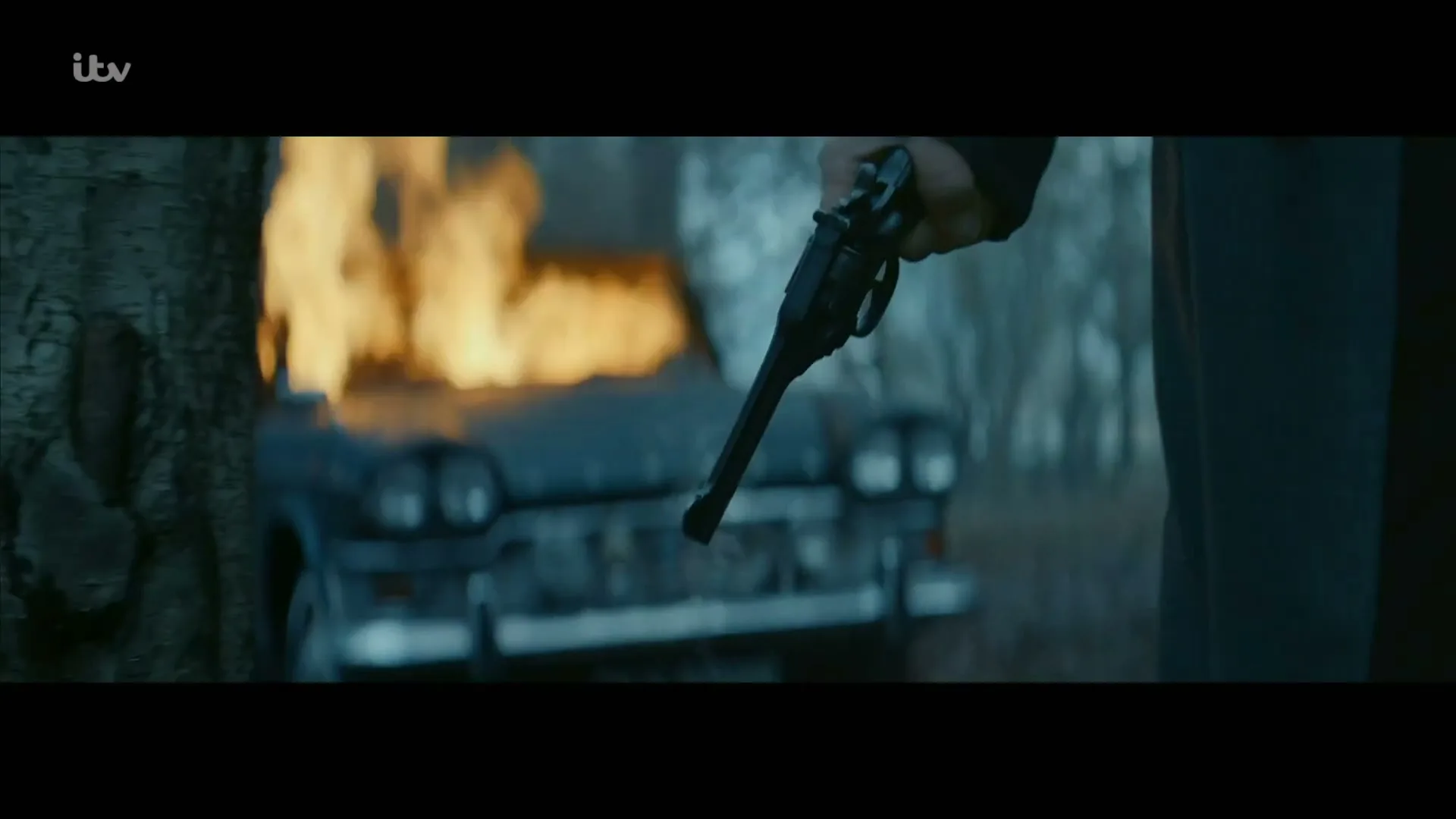 A man in a dark coat holds a smoking revolver, standing in a forest with a burning car in the background.