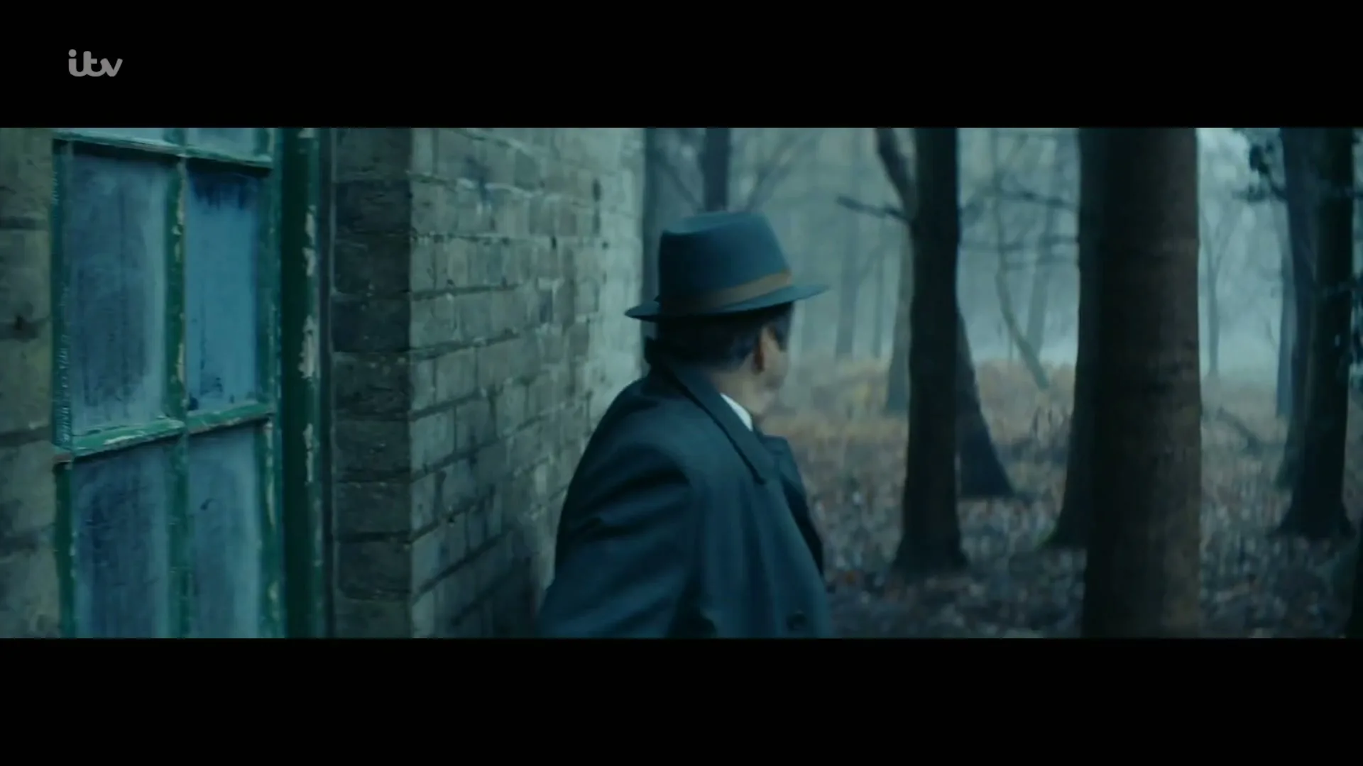 A man in a dark coat and fedora walks past a brick building with broken windows, looking into a misty forest.