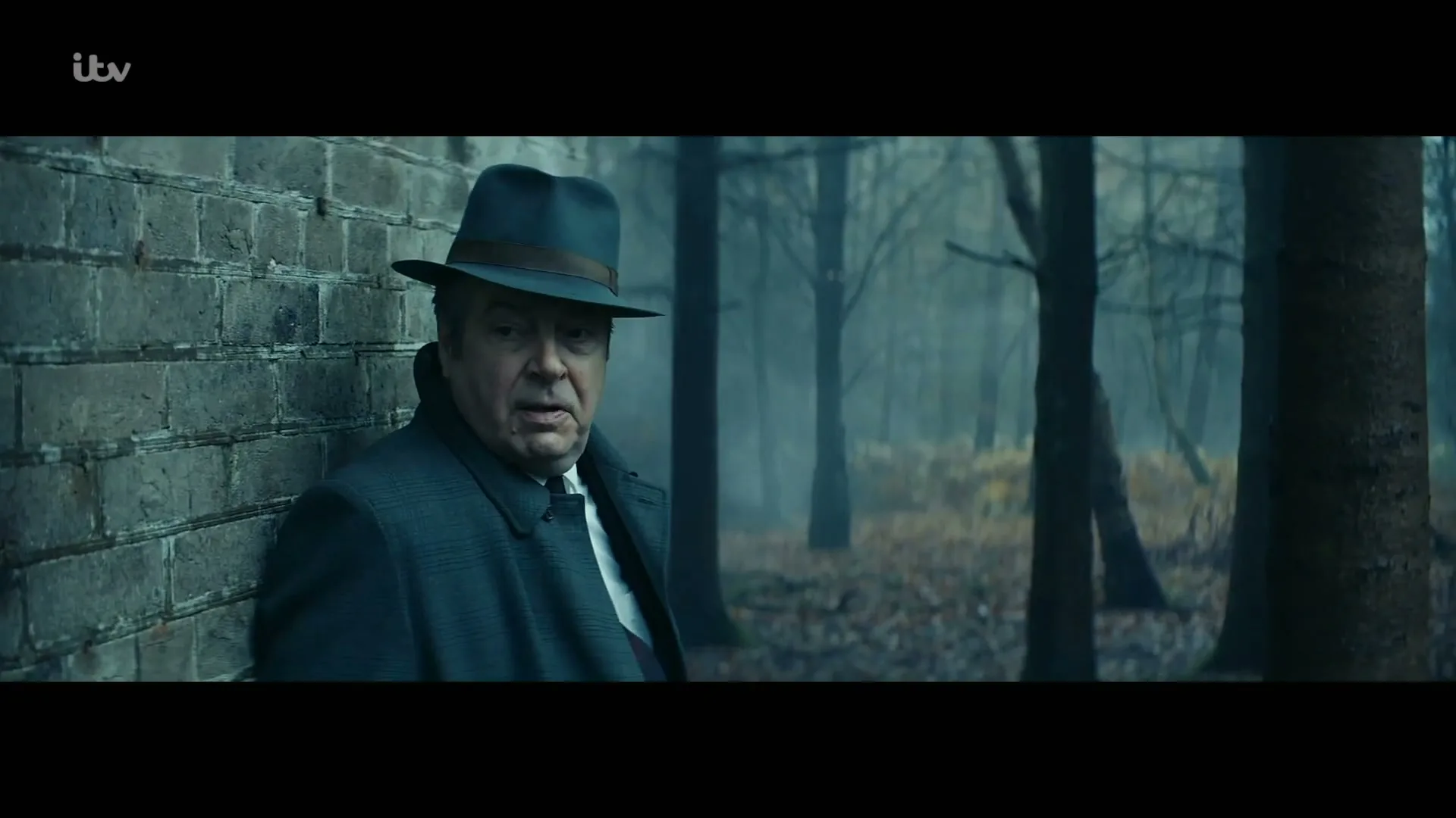 A man in a fedora and trench coat leans against a brick wall in a misty forest, looking around with a worried expression.